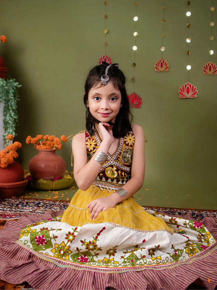 Young girl in a traditional outfit sitting on a decorative mat with a green wall and floral decorations in the background.