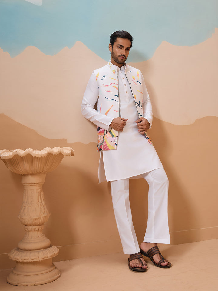 Man in a traditional white sherwani with colorful patterns standing against a beige and blue background.
