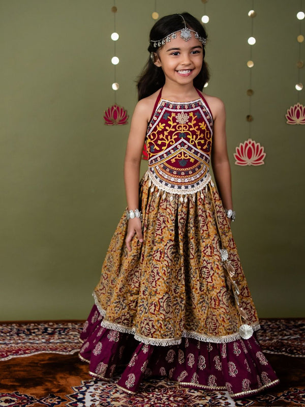 Young girl in a traditional patterned dress standing against a green wall with decorative lights.