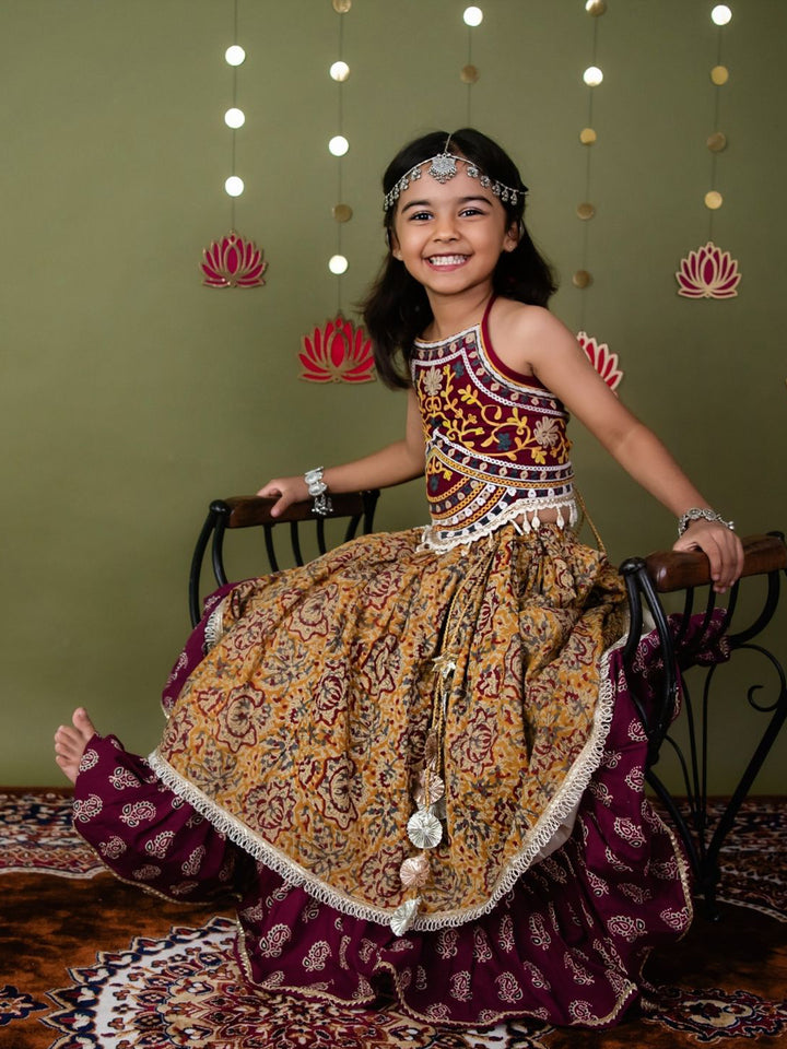 Young girl in a traditional outfit sitting on a chair with decorative lights and flowers in the background.