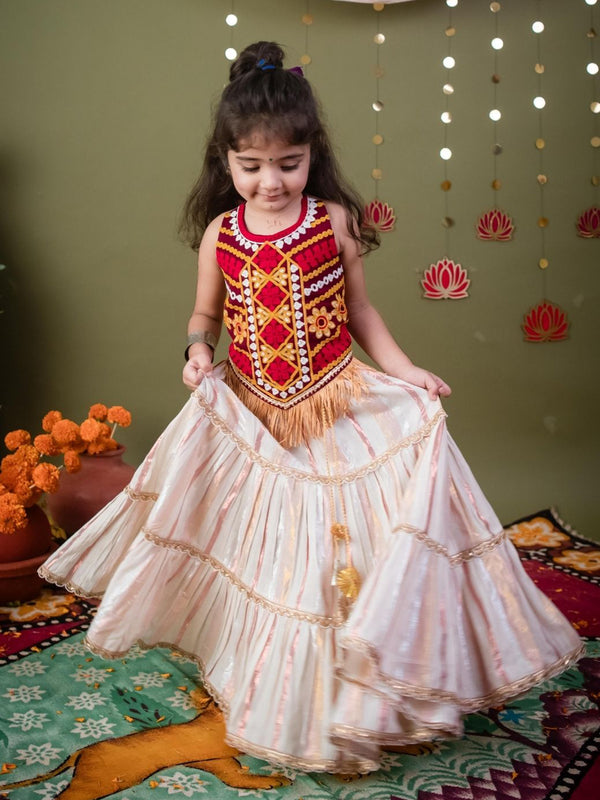 Young girl in a traditional outfit standing on a patterned rug with decorative elements in the background.