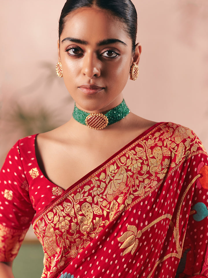 Woman wearing a red saree with gold patterns and a green necklace against a blurred background