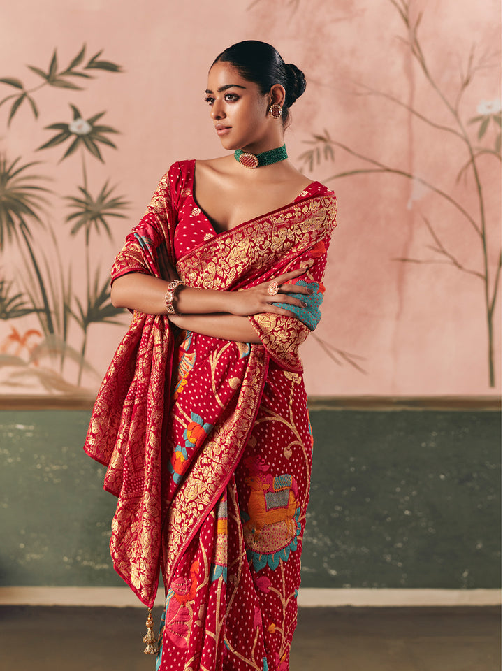 Woman wearing a red saree with intricate patterns against a pink and green background
