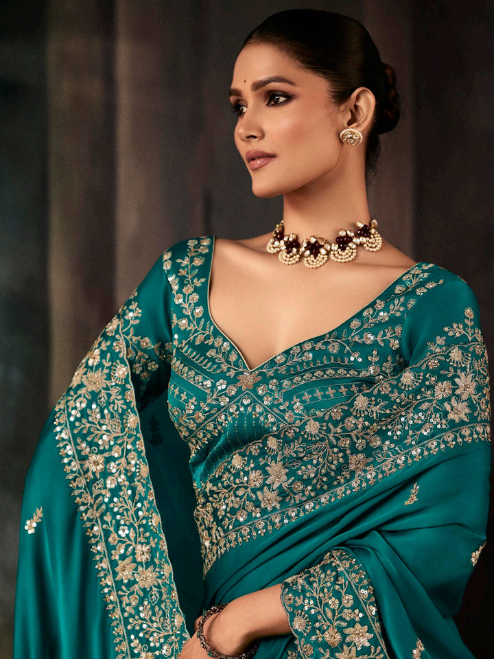 Woman wearing a teal saree with gold embroidery against a dark background