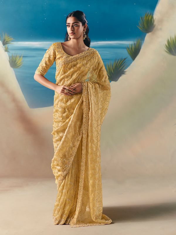 Woman in a yellow saree standing against a scenic background with water and plants.