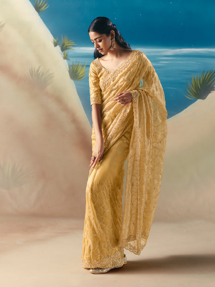 Woman in a yellow saree standing against a scenic background with blue sky and greenery.