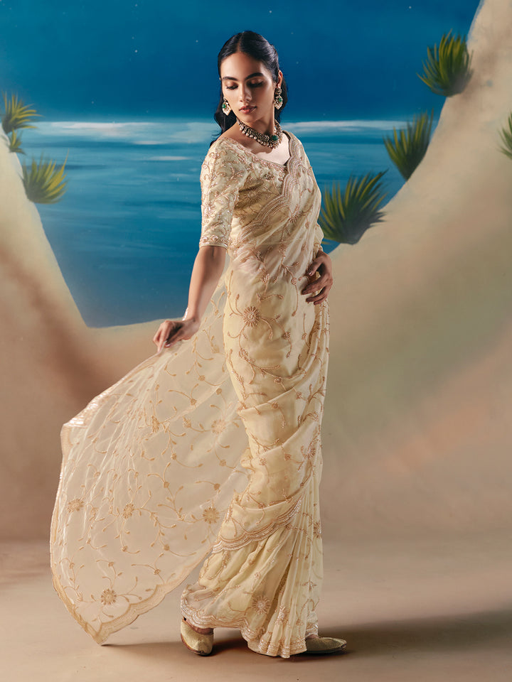 Woman in a cream saree standing against a scenic backdrop with water and plants.