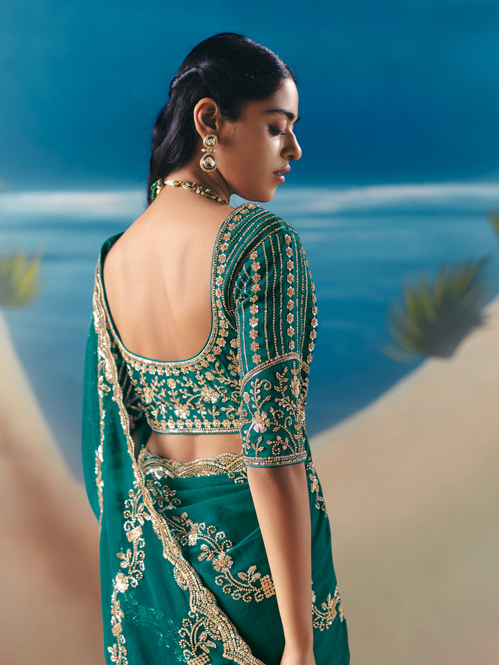 Woman in a green saree with gold embroidery standing by a body of water.