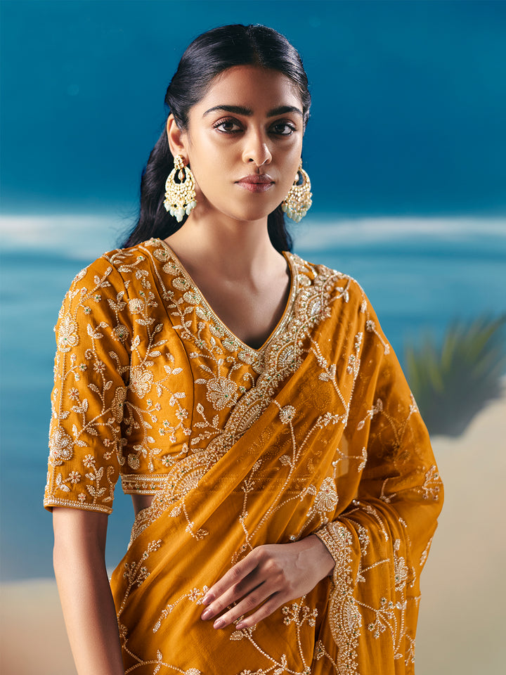 Woman wearing a mustard yellow saree with intricate patterns against a blue and green background