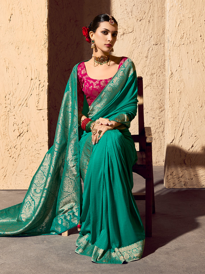Woman in a teal saree with gold patterns sitting against a textured wall.