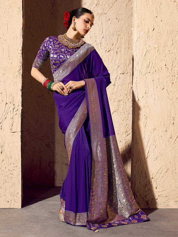 Woman wearing a purple saree with gold border against a textured wall.