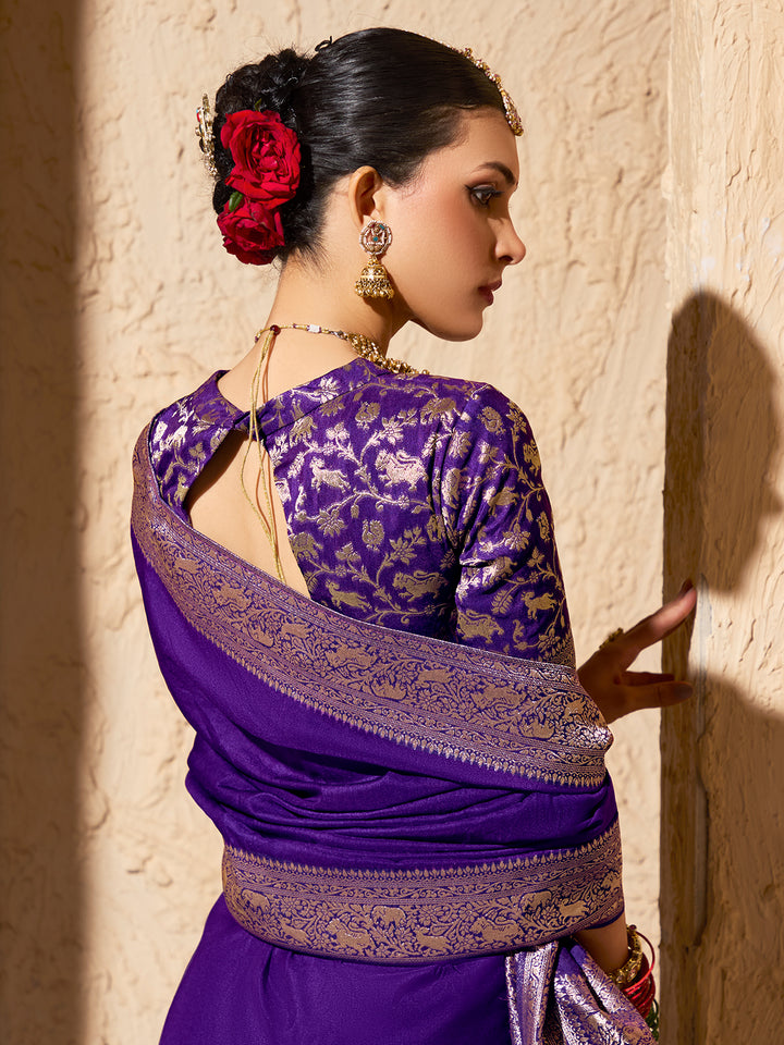 Woman wearing a purple saree with gold patterns against a beige wall.