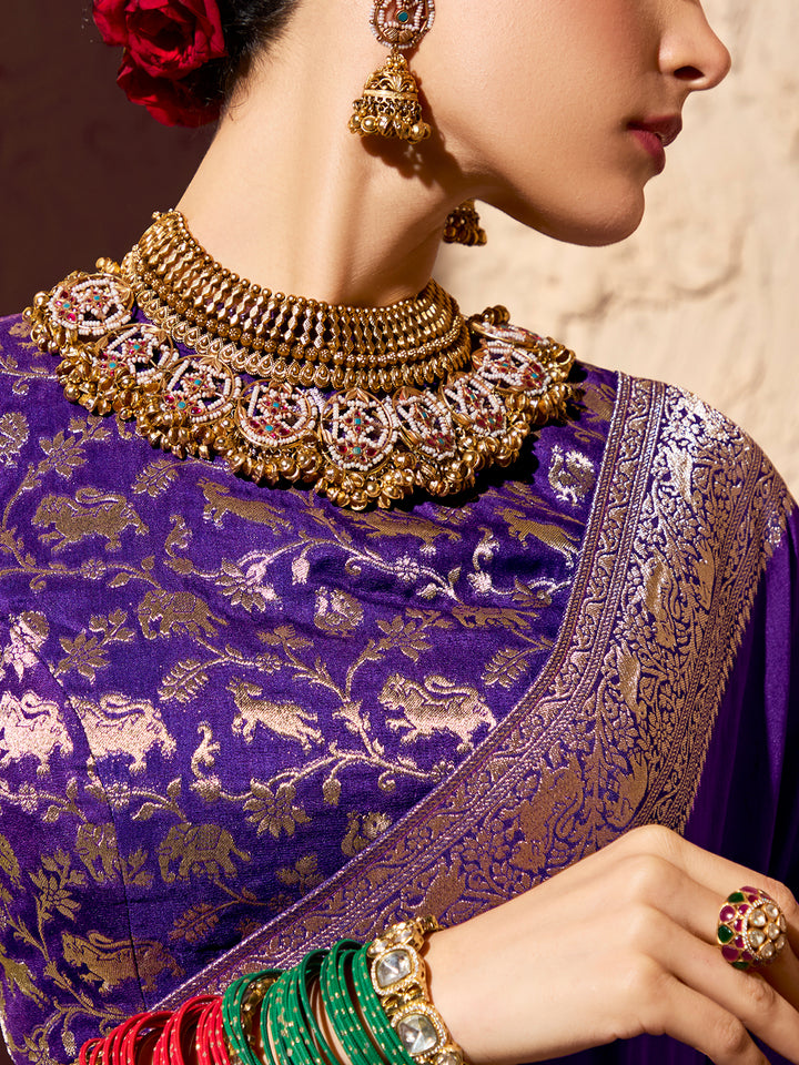 Woman wearing a purple saree with gold embroidery, gold necklace, earrings, and green bangles.