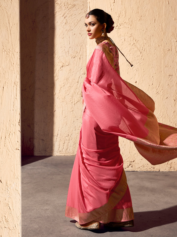 Woman in a pink saree with a gold border against a beige wall.