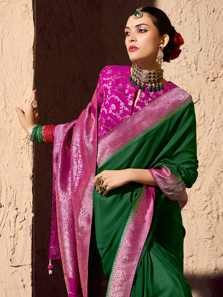 Woman wearing a green and pink saree with jewelry against a textured wall.