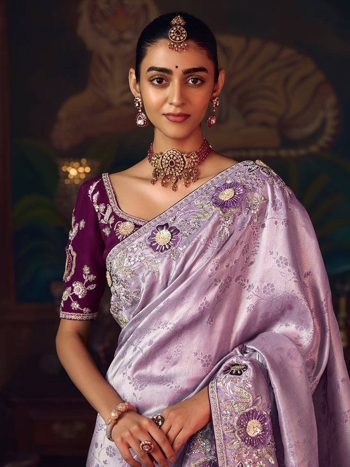 Woman wearing a traditional purple and lavender saree with intricate designs and jewelry.