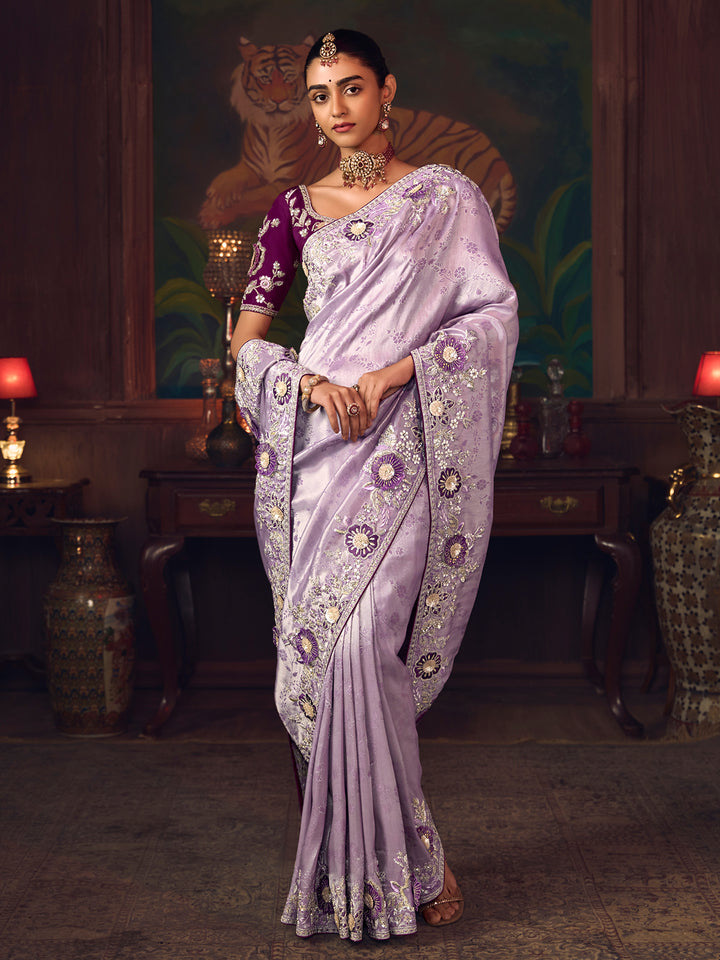 Woman in a lavender saree with intricate patterns in a decorative setting.