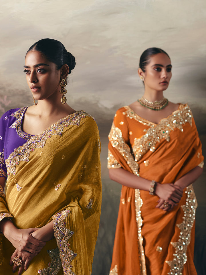 Two women wearing traditional sarees with gold embroidery against a neutral background
