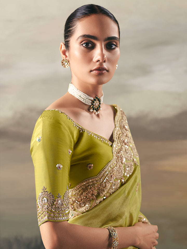 Woman wearing a green saree with gold embroidery against a cloudy sky background