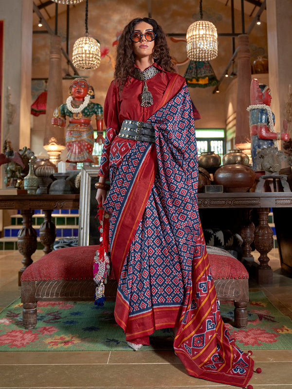 Woman in a red blouse and blue patterned saree standing in an ornate room.