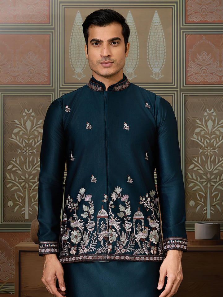 Man wearing a dark blue embroidered traditional outfit against a decorative wall.