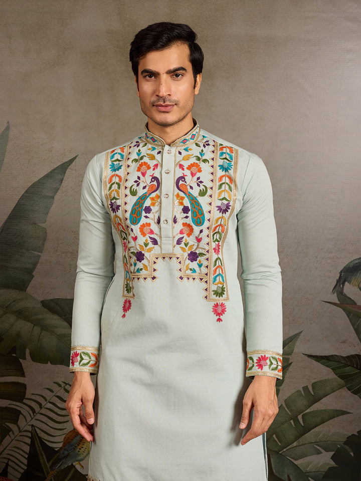 Man wearing a light green kurta with colorful embroidery against a leaf-patterned background