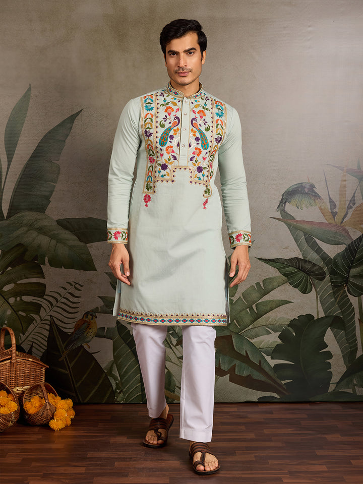 Man wearing a light green embroidered kurta with white pants against a decorative wall.