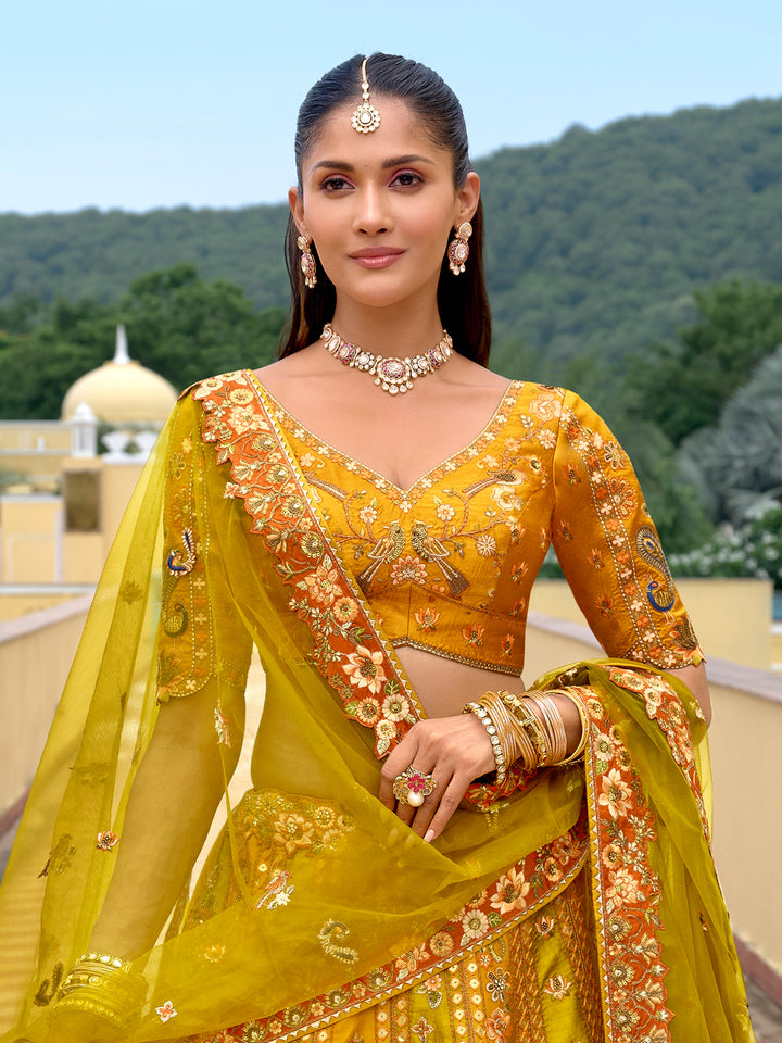 Woman in a yellow traditional outfit with intricate designs, standing outdoors with greenery in the background.