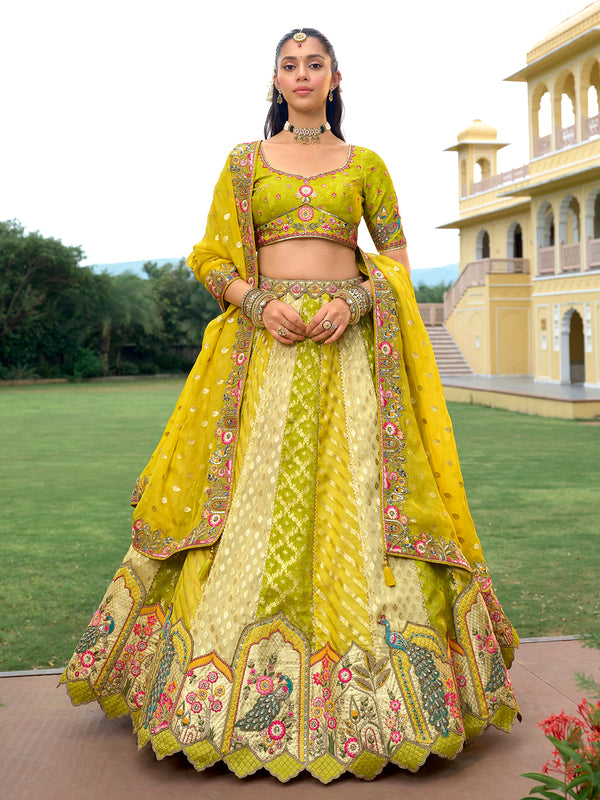 Woman in a yellow and green traditional outfit with intricate patterns, standing in front of a building with greenery.