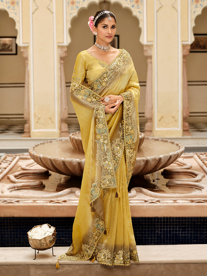 Woman in a yellow saree with intricate patterns standing in an ornate indoor setting.