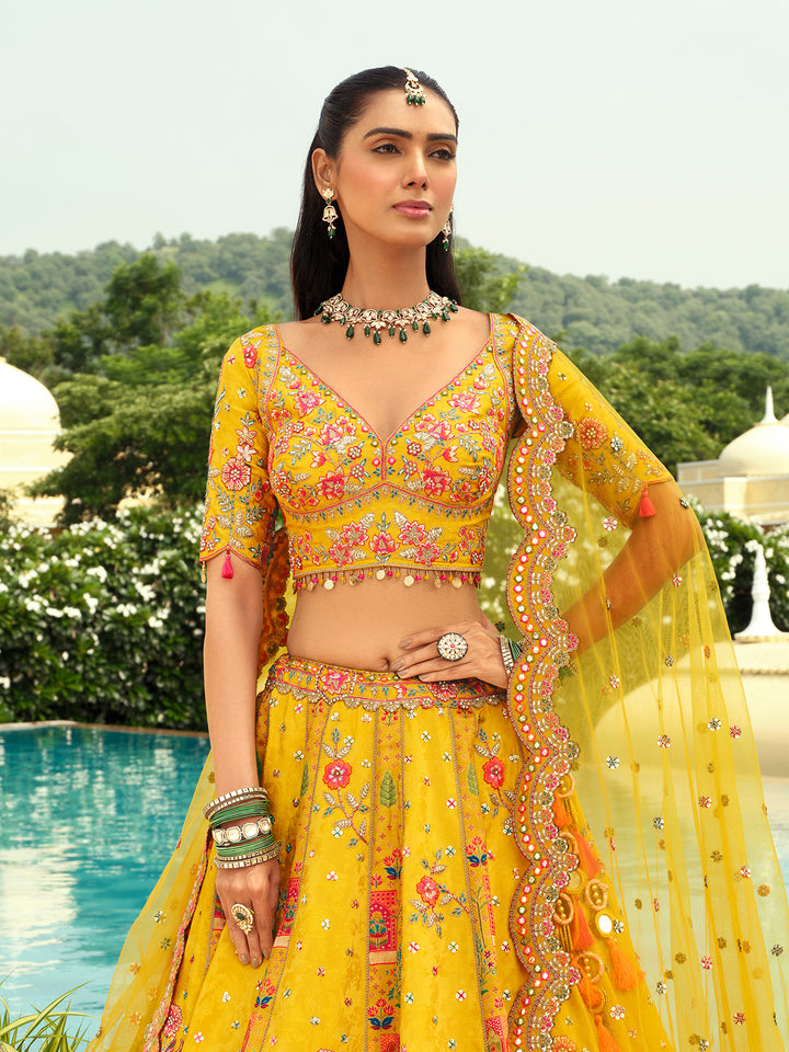 Woman in a yellow traditional outfit with floral patterns, standing outdoors with greenery and a pool in the background.
