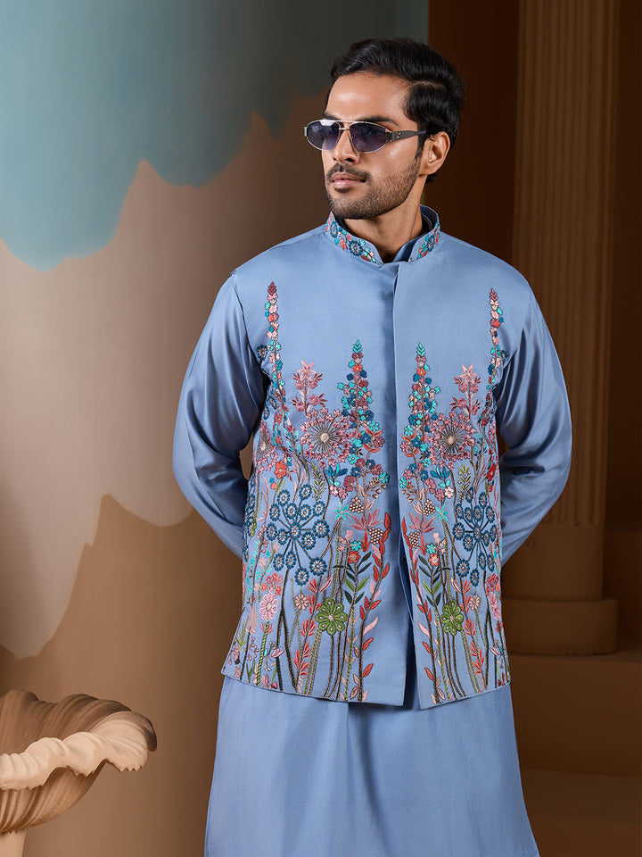 Man wearing a light blue embroidered jacket and pants against a decorative wall.