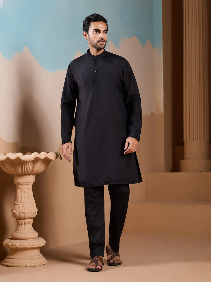 Man wearing a black kurta standing in a decorative indoor setting with a blue wall and stone sculpture.