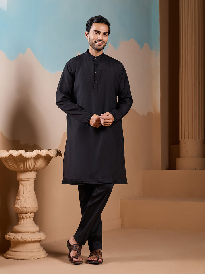 Man wearing a black kurta standing in front of a decorative wall with a blue sky and beige mountains.